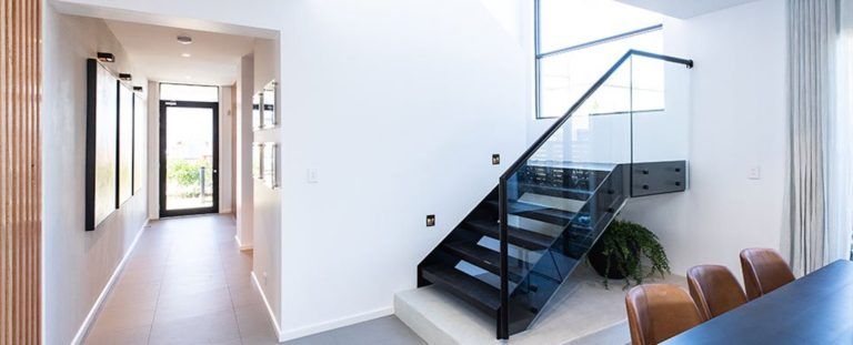 Black steel stairs with glass balustrade and matching black steel l handrail leads into a dining space with a long hallway to the front door.