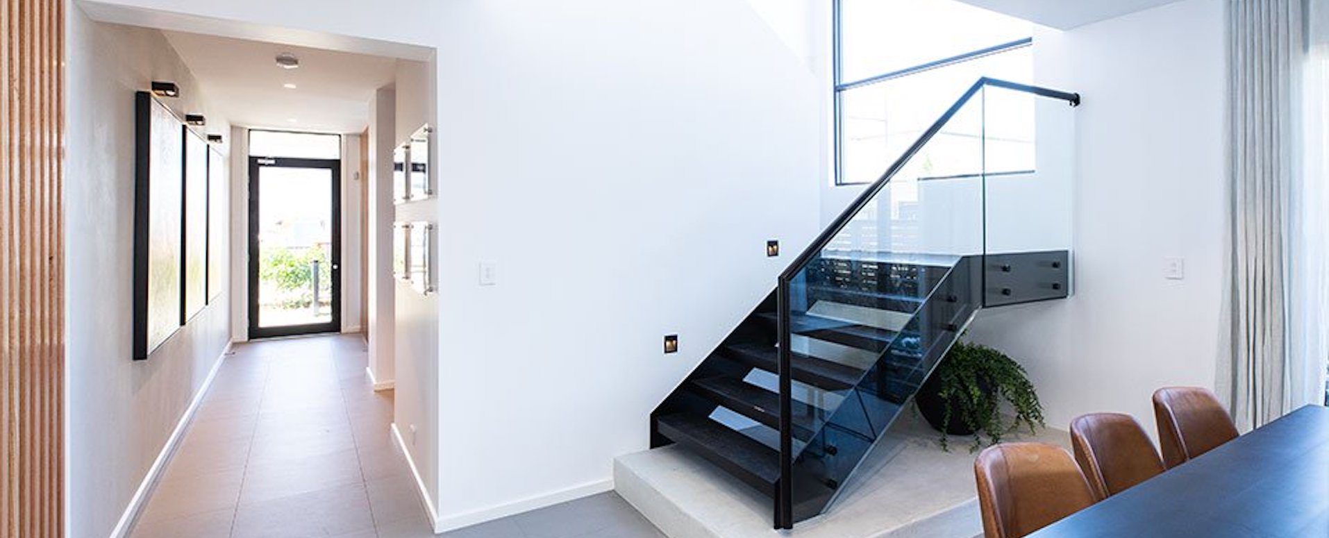 Black steel stairs with glass balustrade and matching black steel l handrail leads into a dining space with a long hallway to the front door.