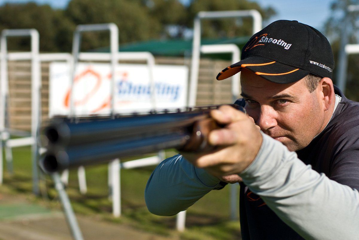 Man wearing a black cap with text "Oz Shooting" and holding a rifle pointed just past the camera. "Oz Shooting" sign blurred in the background