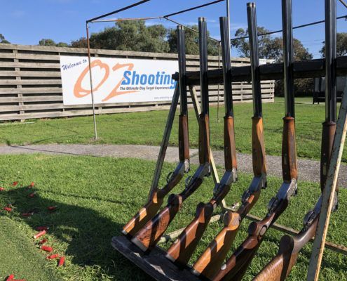Six rifles on a stand with a sign in the background with blue and orange text “Welcome to Oz Shooting the ultimate clay target experience”