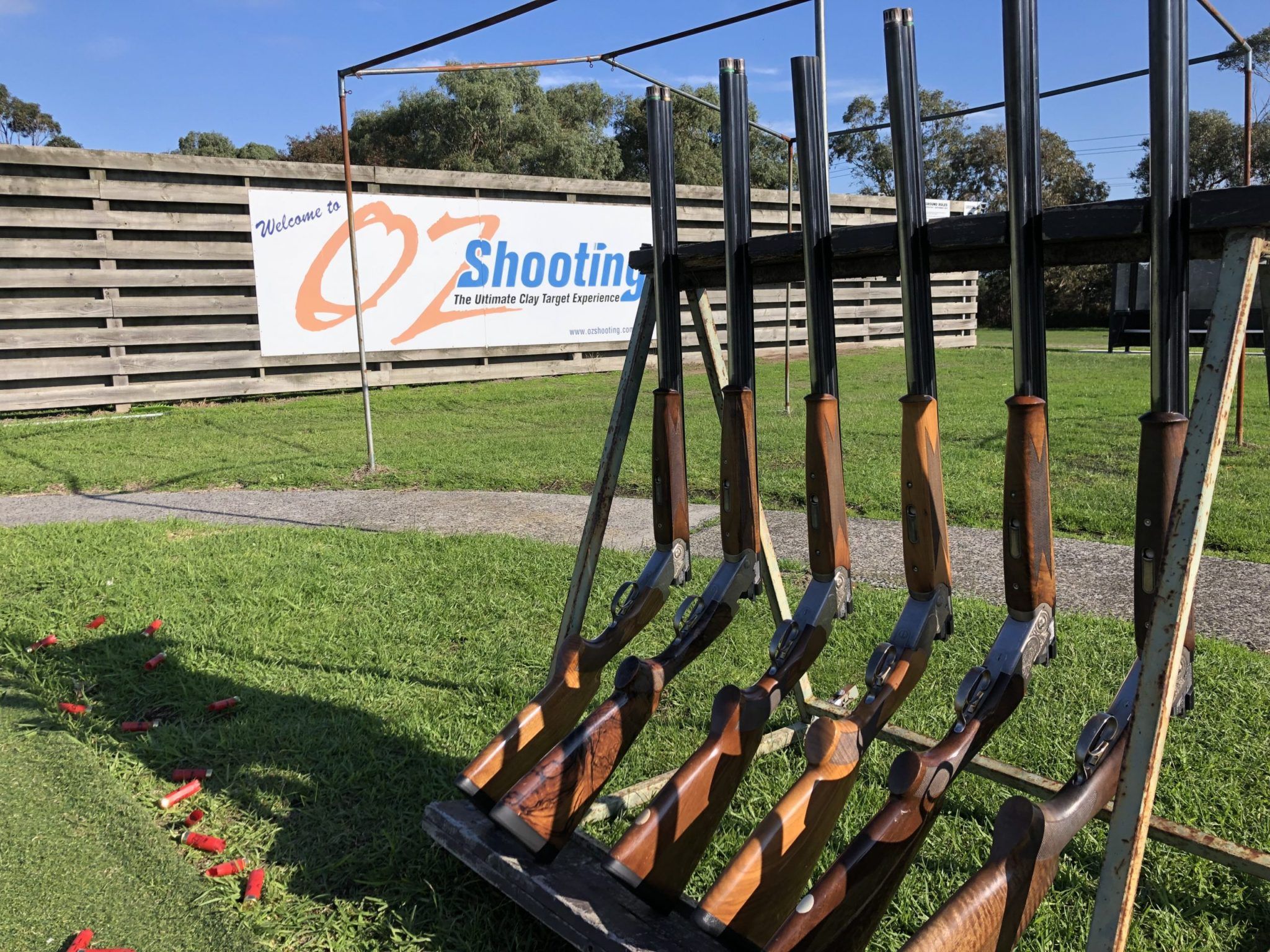 Six rifles on a stand with a sign in the background with blue and orange text “Welcome to Oz Shooting the ultimate clay target experience”