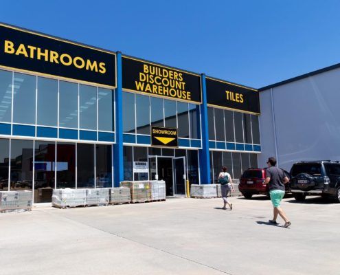 Customers walking across carpark to a big warehouse showroom. Black background with bright yellow text “Builders Discount Warehouse”