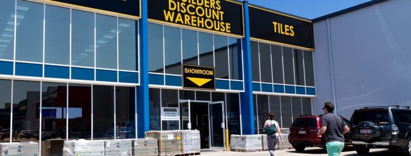Customers walking across carpark to a big warehouse showroom. Black background with bright yellow text “Builders Discount Warehouse”