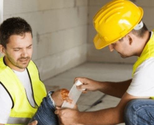 2 employees with high vis and hard hats. One is kneeled down putting a bandage on an injured knee.