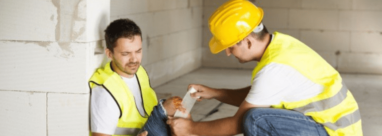2 employees with high vis and hard hats. One is kneeled down putting a bandage on an injured knee.
