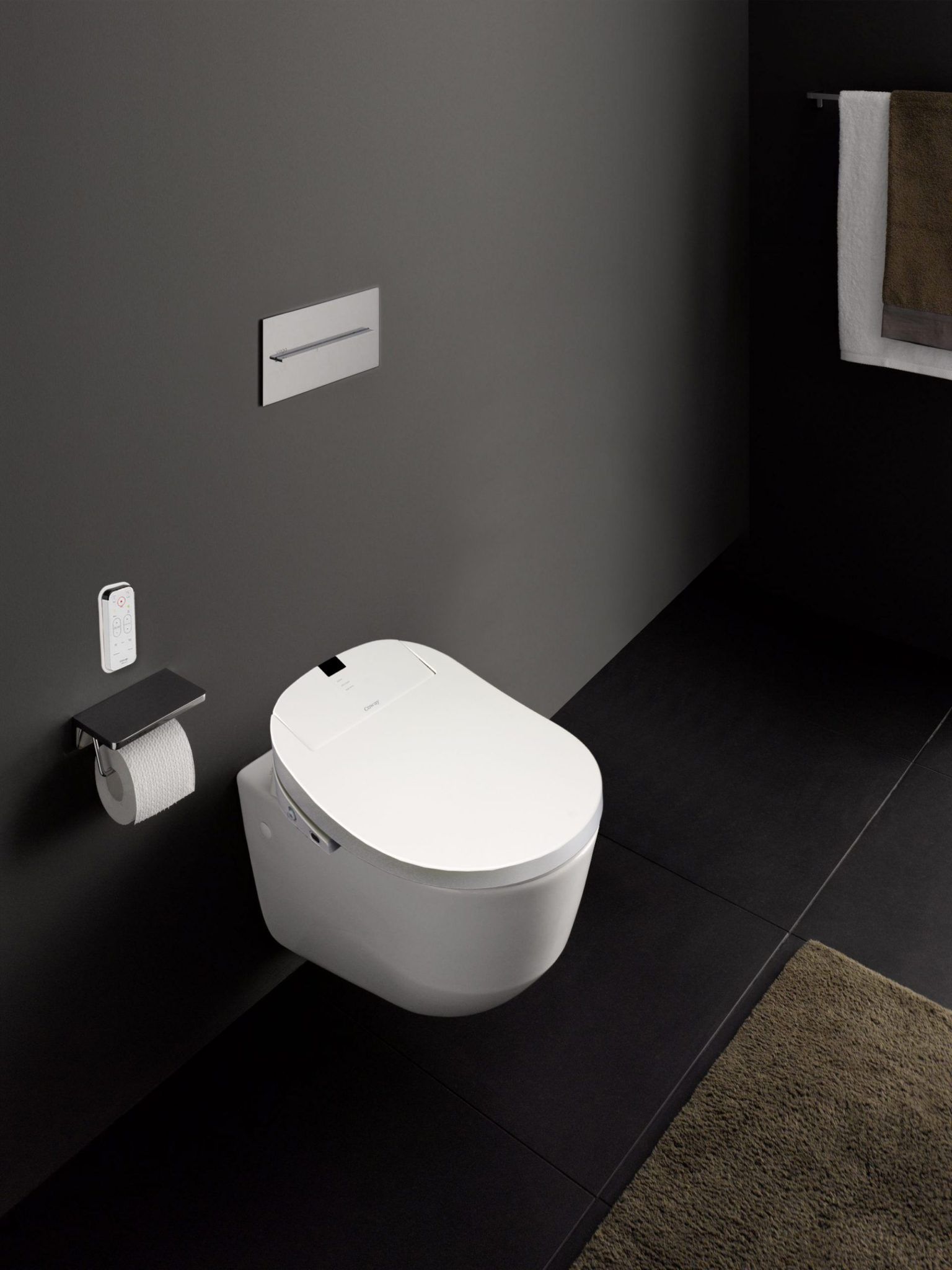 Minimalist and monochrome bathroom with white floating toilet and toilet paper. Black painted walls and black tiles. Brown bath mat and towels.