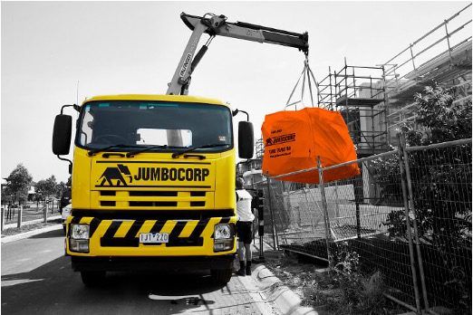 Yellow Jumbocorp truck parked outside a construction site, loads an orange waste bag onto it.