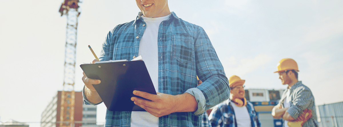 close up of builder holding clipboard on site and workers in background